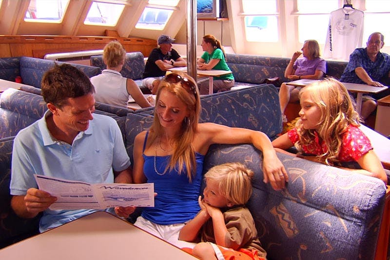 A family sits in a boat&#39;s lounge area, with the parents looking at a menu and the child resting his head on his mother&#39;s arm. Other passengers are visible in the background.