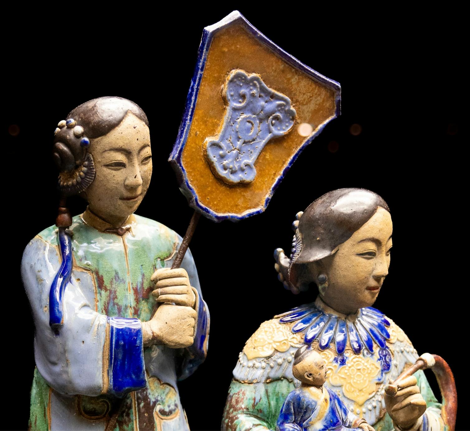 Två färgglada keramiska figurer, en som håller en sköld och den andra som håller en docka, visas mot en mörk bakgrund.