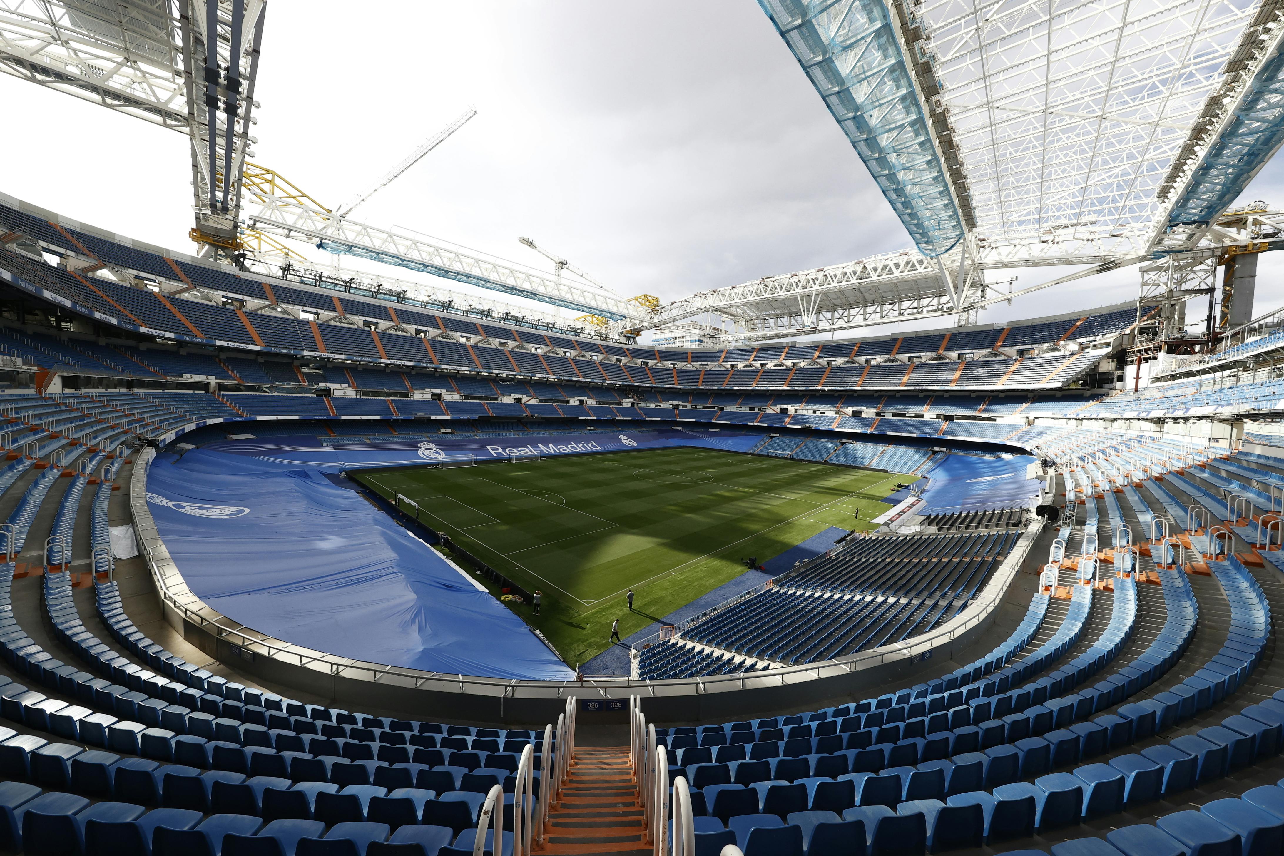 Empty football stadium with blue seats and a green pitch under construction cranes and a partially retracted roof.