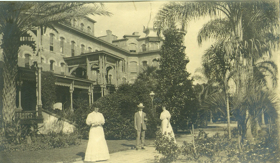 Three people in period clothing stand in a garden in front of a large, ornate building surrounded by trees and foliage.