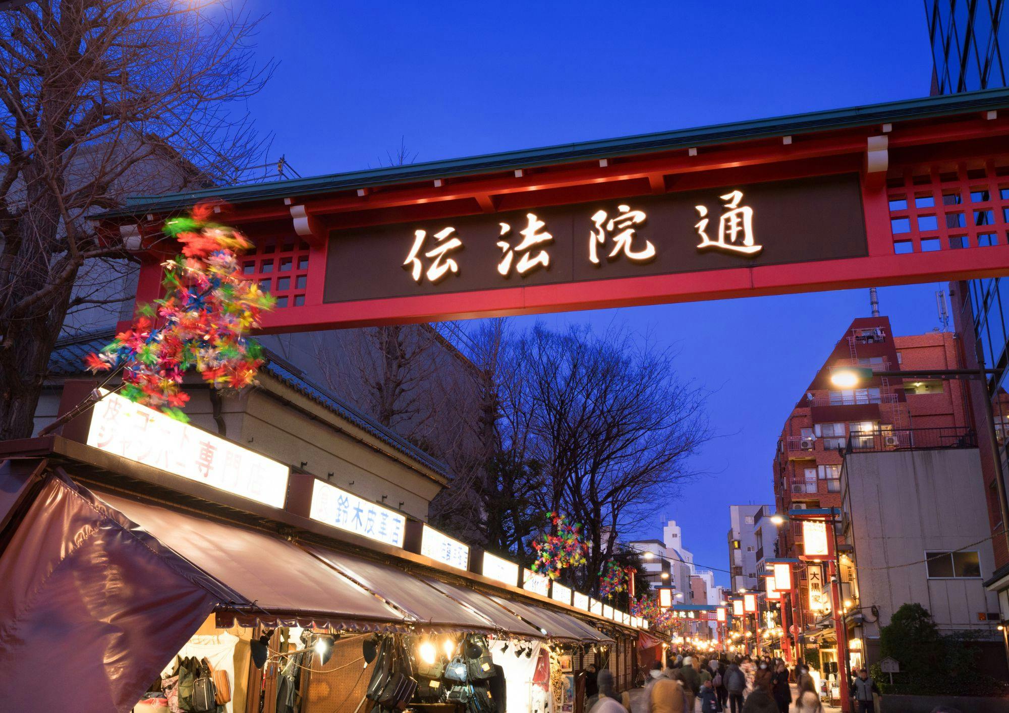 A view of a Japanese market street at dusk, with a sign featuring Japanese characters and shops lining both sides.