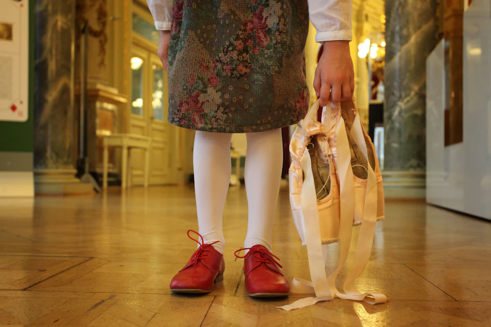 A child standing indoors holding ballet slippers, wearing white tights, red shoes, and a floral-patterned dress.