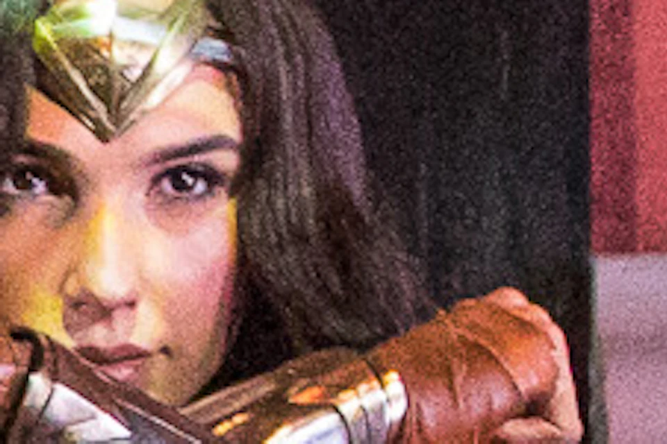 Four people are posing around a Wonder Woman statue, mimicking her crossed-arm stance, in a room with orange walls and shadows.