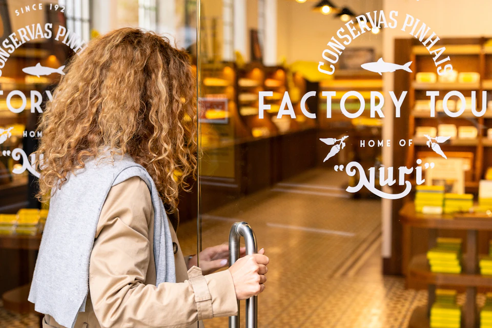 A person with curly hair opens a glass door to a store with visible signage and a warmly lit interior featuring shelves.