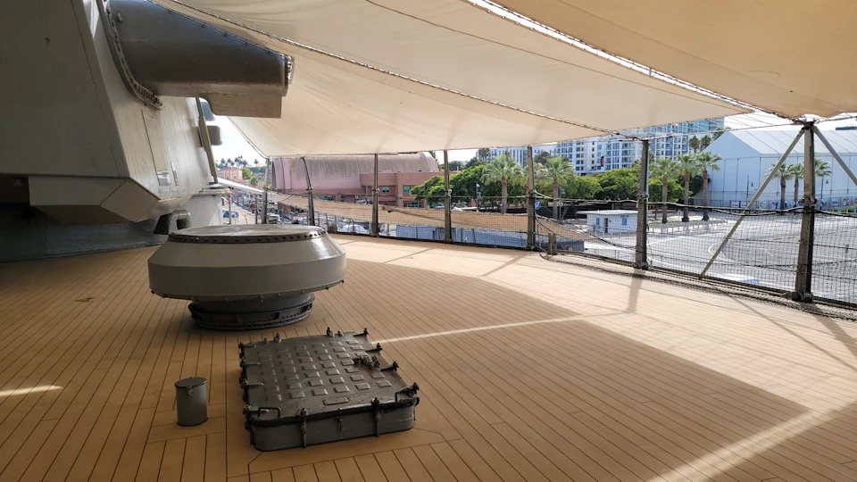 A ship's deck with covered shade, a hatch, and mechanical equipment; buildings and trees are visible in the background.