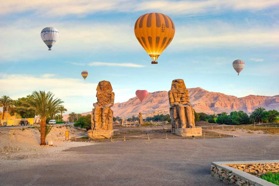 Hot air balloons floating over ancient stone statues in an open area with mountains and greenery in the background.