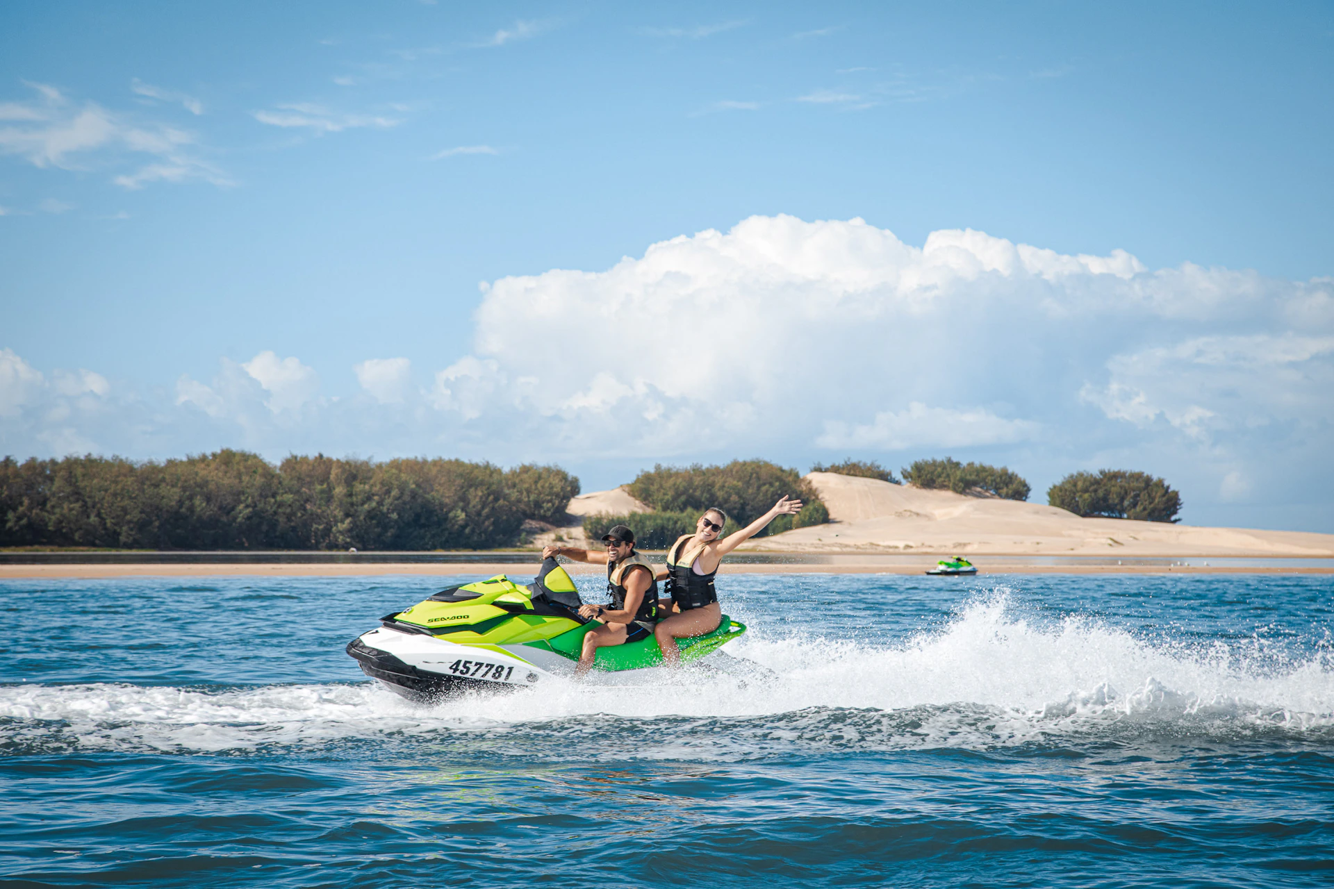 Two people riding a green jet ski on a sunny day with sand dunes and trees in the background.