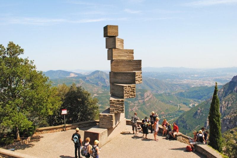 People gather near a large, stacked stone sculpture overlooking a scenic mountain landscape under a clear blue sky.