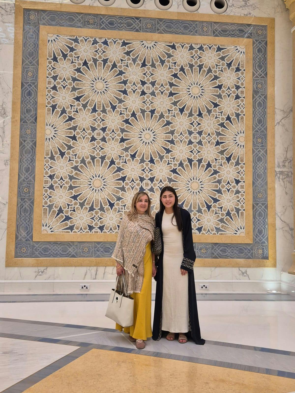 Two women standing in front of an ornate wall decorated with a geometric floral pattern. One wears a yellow outfit, the other a white dress.