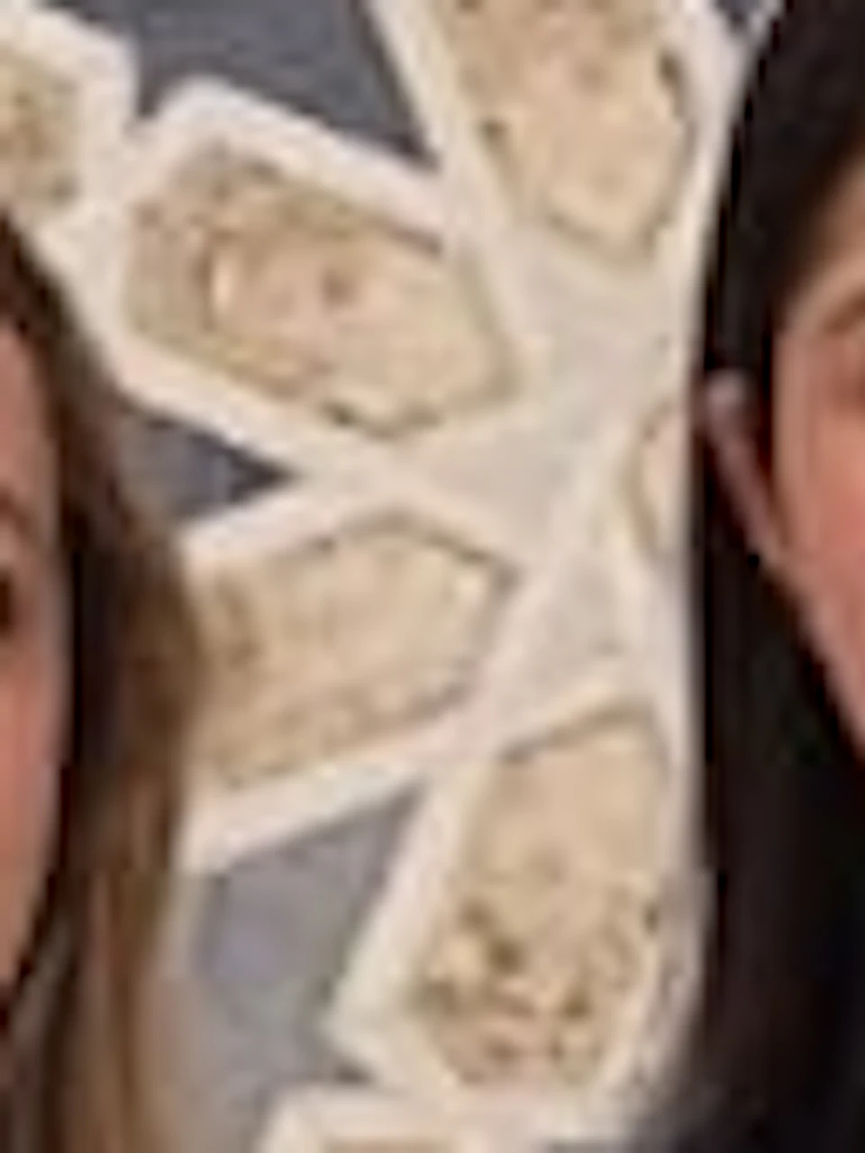 Two women standing in front of an ornate wall decorated with a geometric floral pattern. One wears a yellow outfit, the other a white dress.