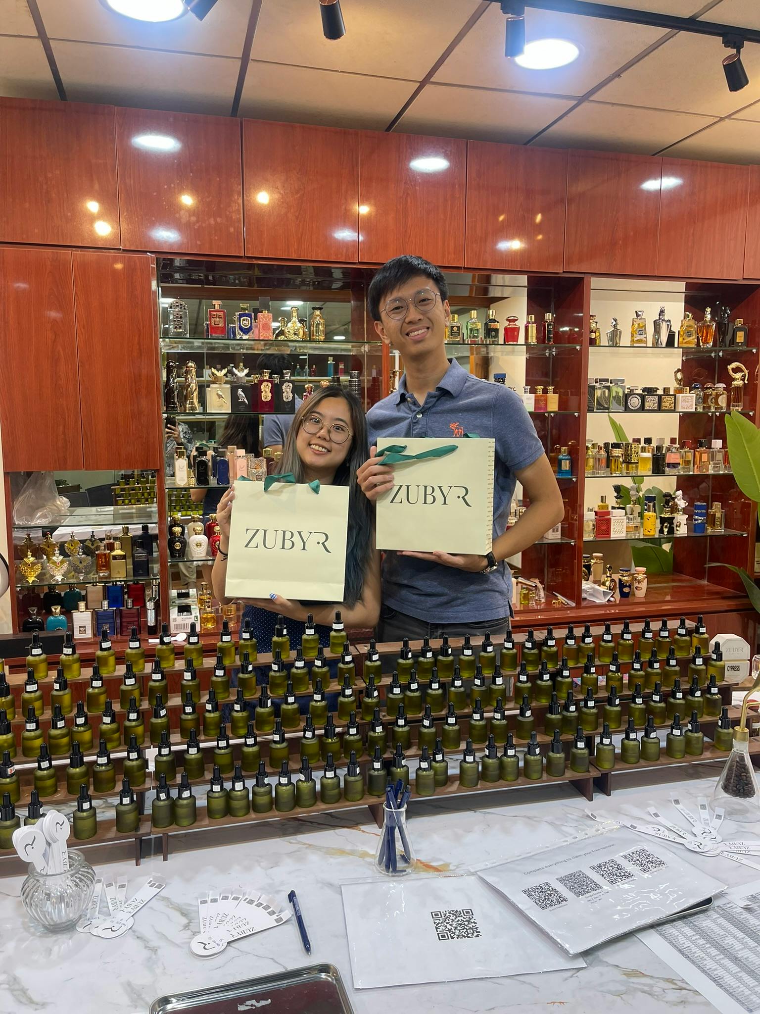 Two people smiling and holding shopping bags labeled "Zubyr" in a store with shelves of perfume bottles.