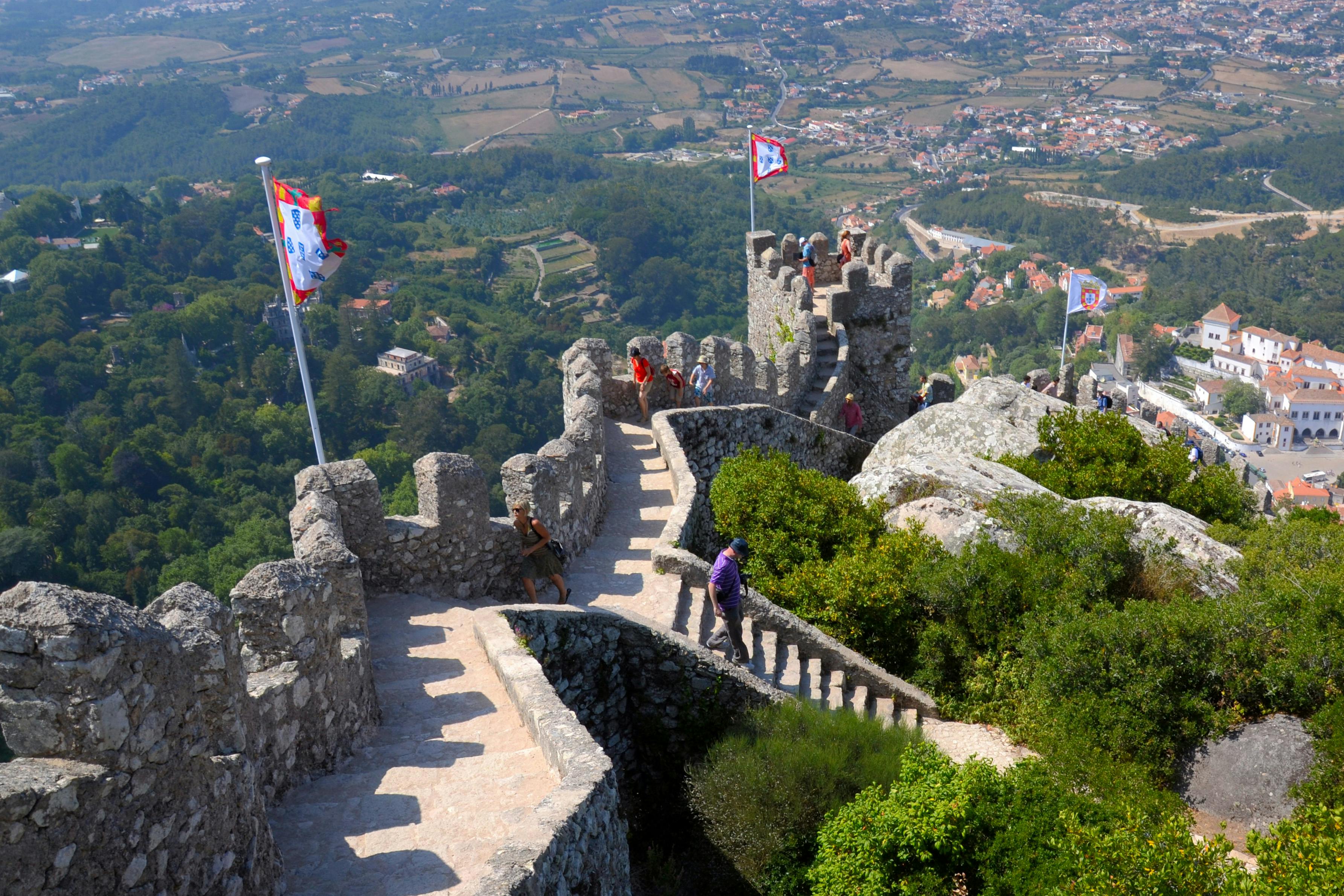 Des touristes marchent le long d'un mur de forteresse en pierre, drapeaux flottants, surplombant une vallée verdoyante et une ville lointaine.