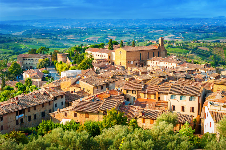 Aerial view of a medieval town with terracotta-roofed buildings, a prominent stone church, and surrounding lush green countryside.
