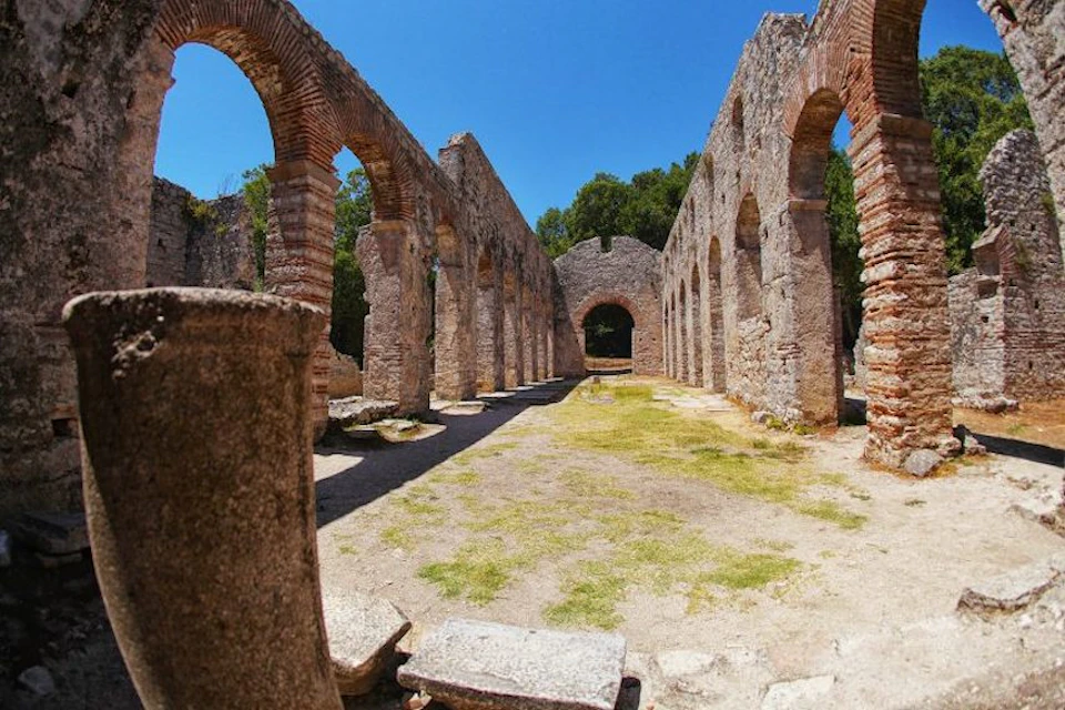 Ancient stone ruins with tall arches and columns under a clear blue sky. Some vegetation visible in the background.