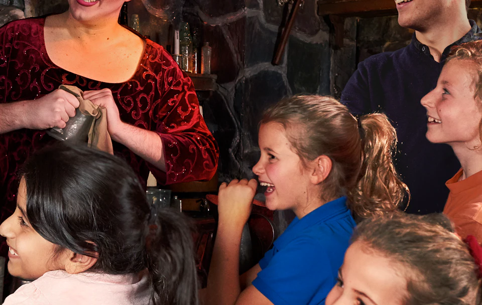A woman in a red dress entertains a group of smiling children and an adult in a dimly lit room filled with various objects on shelves.