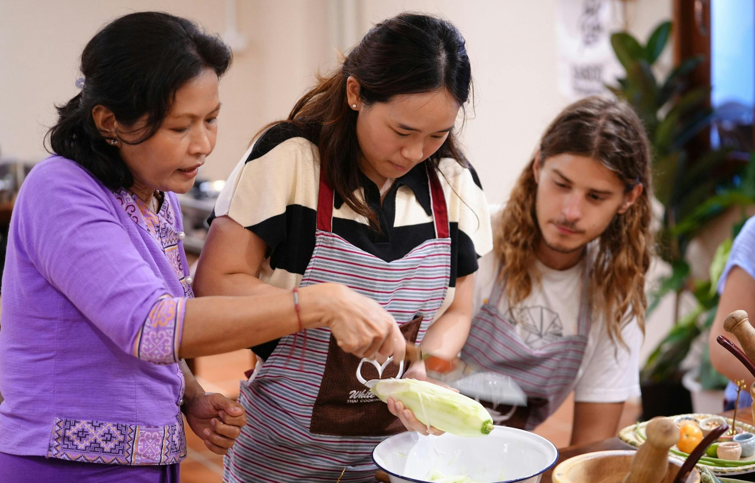 Three people are cooking together, with one holding a vegetable, another providing guidance, and a third observing attentively.