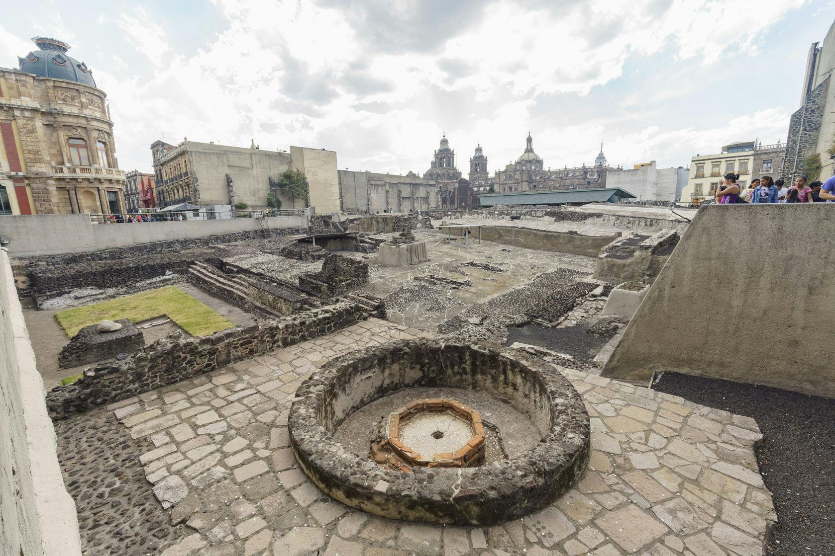 Ruinas de antiguas estructuras de piedra en una zona urbana, con edificios históricos al fondo y un grupo de personas observando.