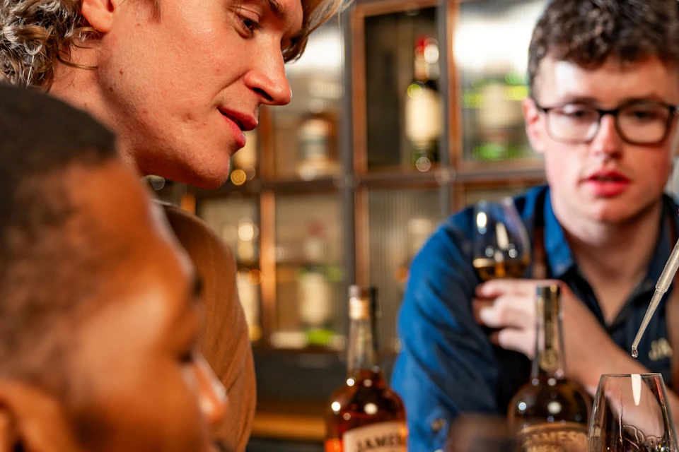 A group of people at a bar counter sampling whiskey, with bottles displayed on shelves in the background.