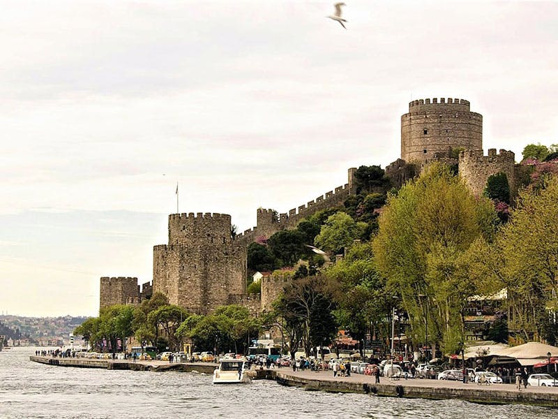 Fortezza di pietra con torri cilindriche su una collina, circondata da alberi, con persone e barche sul lungomare.