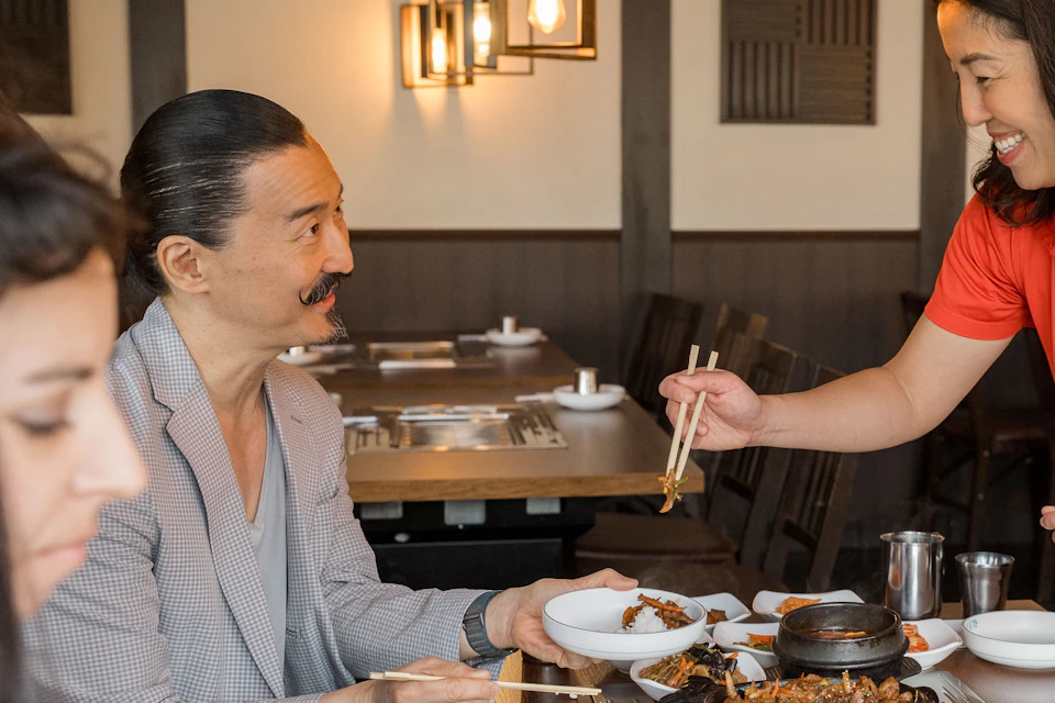 Vancouver Foodie Tour Guide in a bright red shirt serving korean barbeque to a young male with a distinguished mustache.