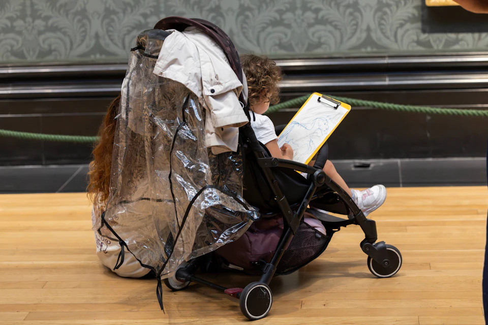 A child in a stroller draws on a clipboard, covered by a transparent rain cover.
