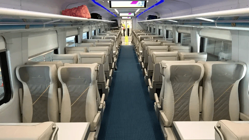 Interior of a modern train carriage with gray seats, blue carpet, and overhead luggage racks. A door is visible at the end.