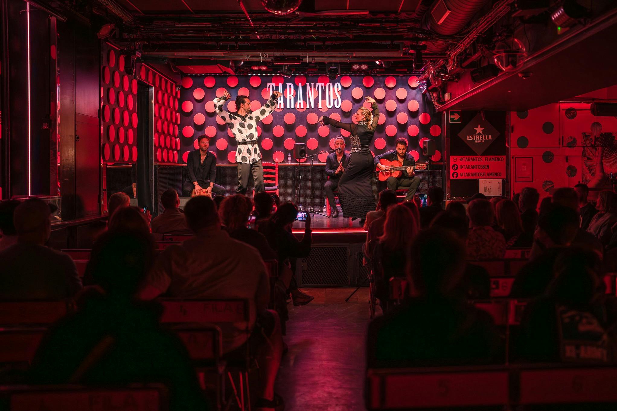 Intèrprets a l'escenari d'un espectacle de flamenc amb un públic assegut en un local poc il·luminat amb decoració de lunars vermells.
