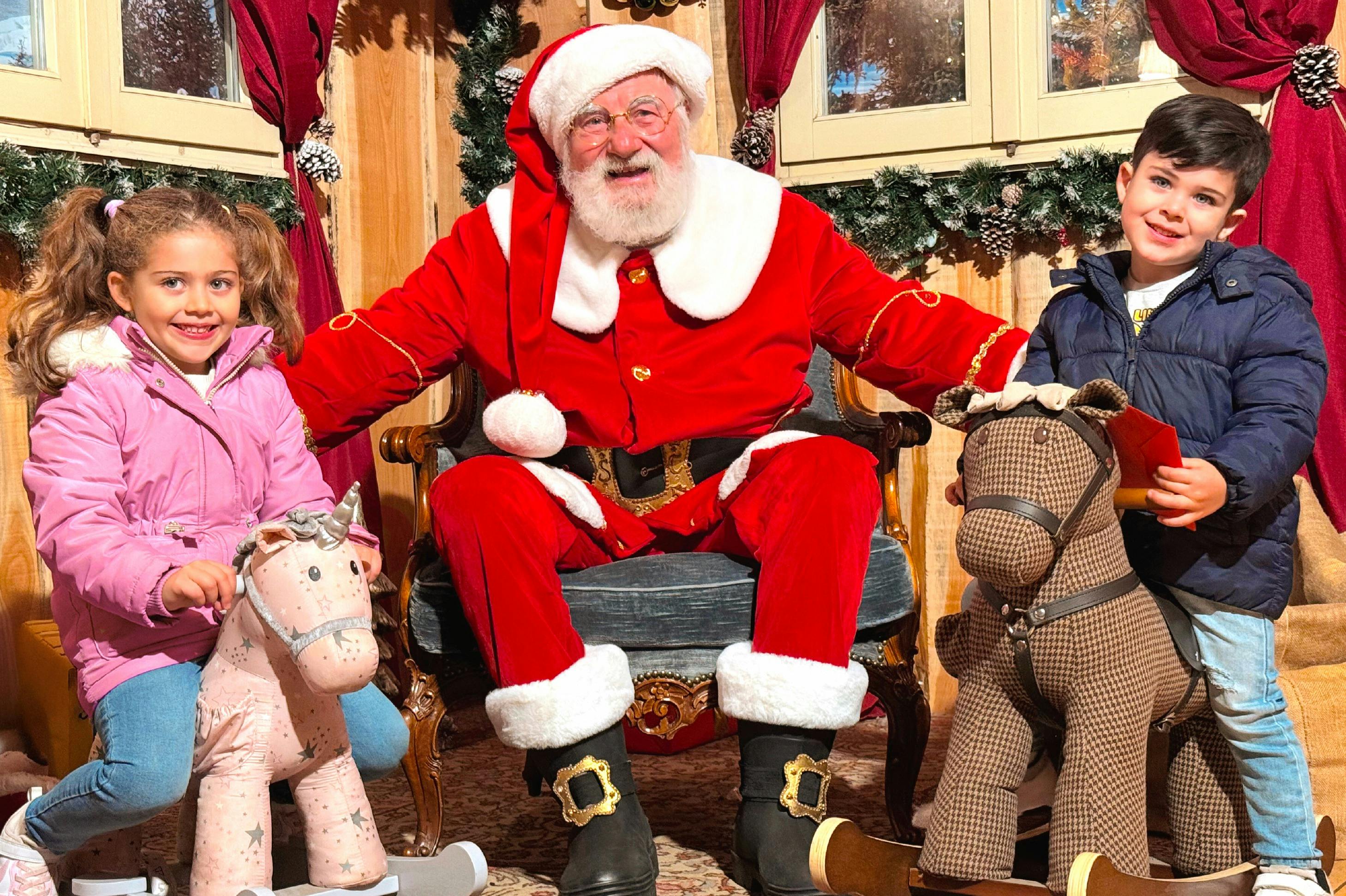 Ένα μικρό κορίτσι με ροζ παλτό κάθεται σε ένα κουνιστό άλογο δίπλα στον Άγιο Βασίλη σε μια καρέκλα μέσα σε ένα εορταστικό δωμάτιο με γιορτινή διακόσμηση.