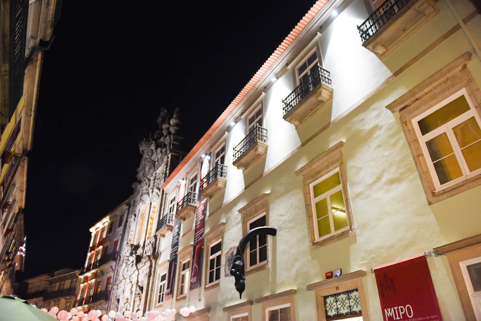 Illuminated multi-story building with balconies, a sculpture attached to the wall, and pink balloons below, all under a night sky.