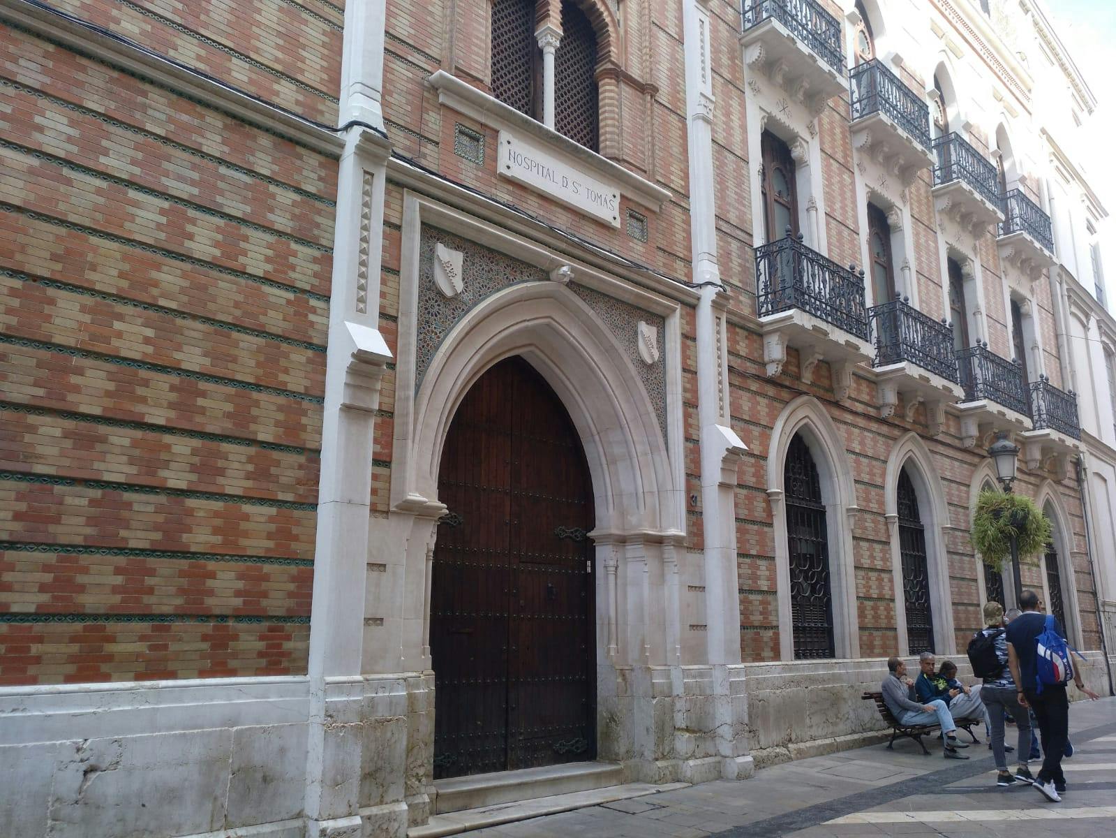 Eine große gewölbte Holztür in einer verschnörkelten Backsteinfassade mit der Aufschrift "Hospital S. Juan" darüber. Menschen sitzen auf einer Bank in der Nähe.