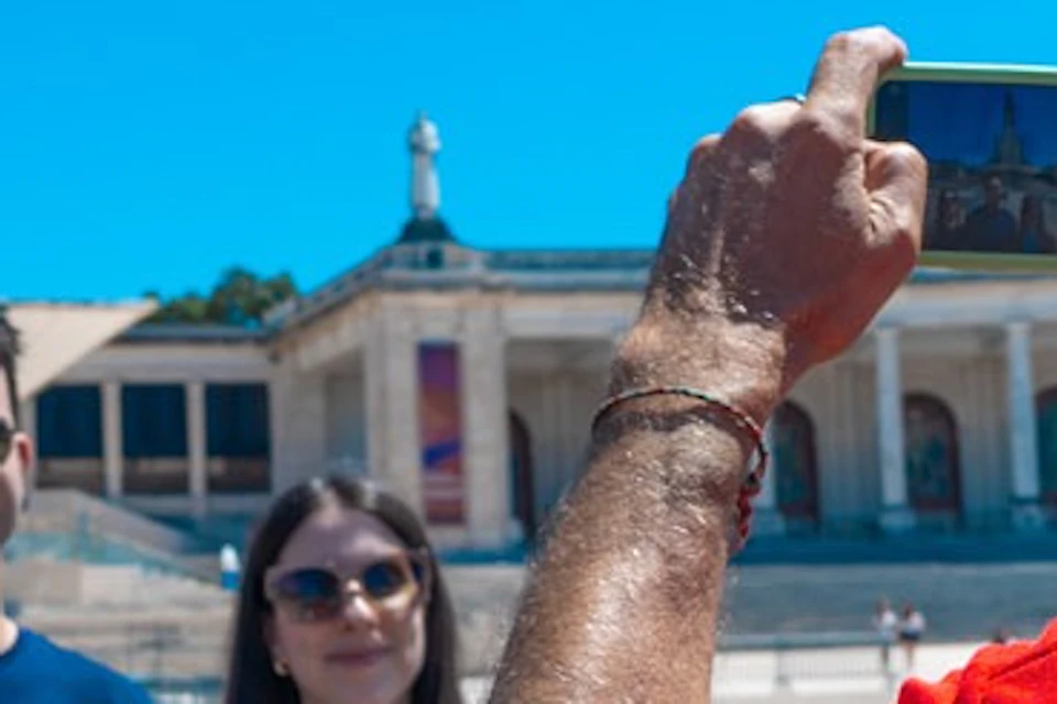 A man in a red shirt takes a photo of three people posing in front of a historic building under a clear blue sky.
