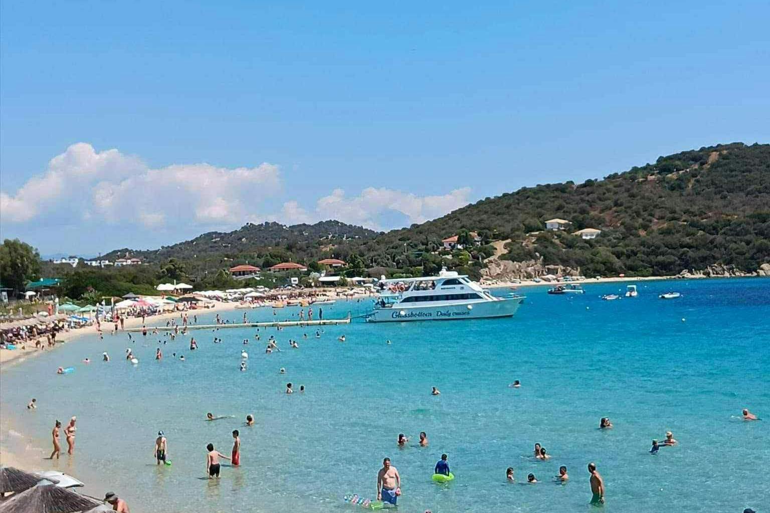 Een druk strand met mensen die zwemmen in het helderblauwe water. Een boot ligt voor anker bij de kust. Heuvels zijn op de achtergrond.
