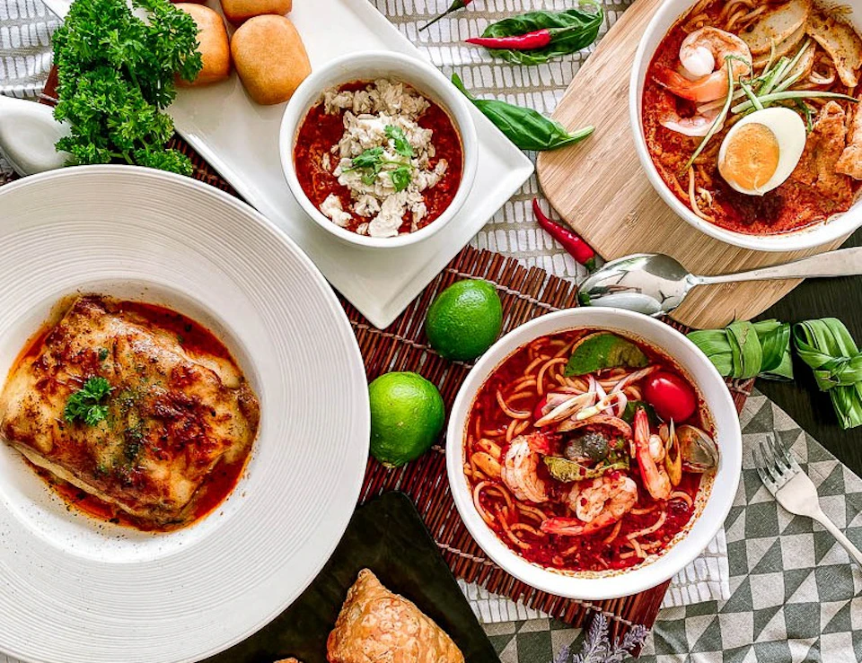 A variety of dishes including soups, bread, limes, and herbs are arranged on a table with vibrant presentation.