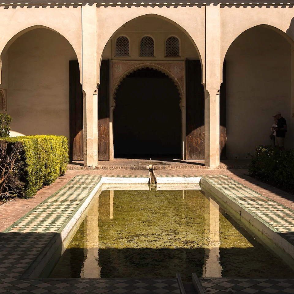 A courtyard with an arched entryway, a reflecting pool, and hedges; a person stands in the background near the shadows.
