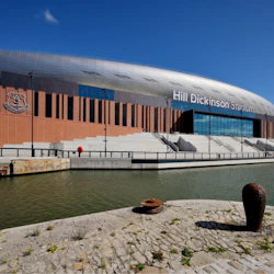 En modern arena märkt "Hill Dickinson Stadium" med en kanal i förgrunden, under en klarblå himmel.