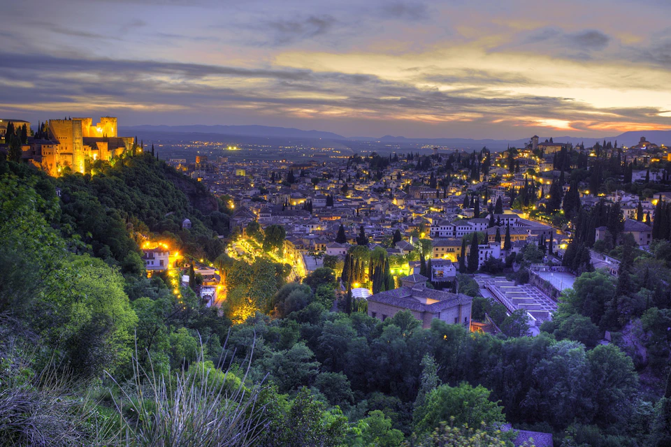 Aerial view of a cityscape at dusk with illuminated buildings, trees, and a cloudy sky.