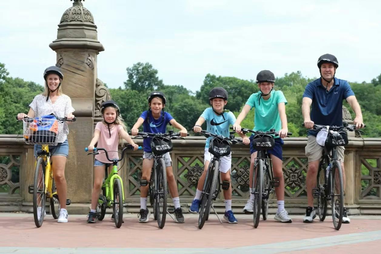 A group of five people, including children, standing with bicycles on a decorative stone bridge on a sunny day.