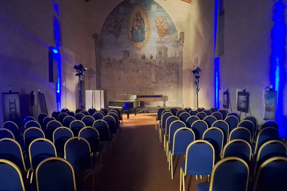 An empty auditorium with rows of blue chairs facing a stage featuring a grand piano and a frescoed wall in the background.
