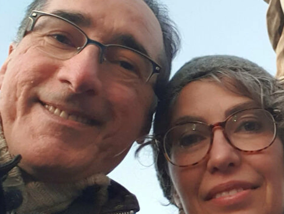Two people wearing glasses pose for a selfie in front of a stone statue with detailed carvings.