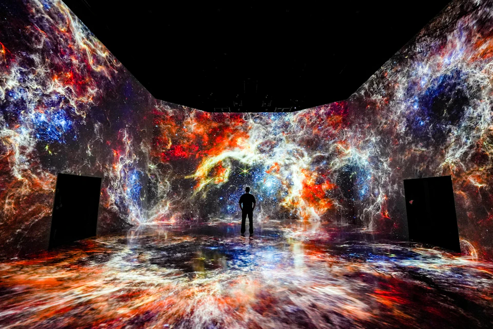 A person stands in a room surrounded by vivid, colorful cosmic visuals projected on the walls and floor.