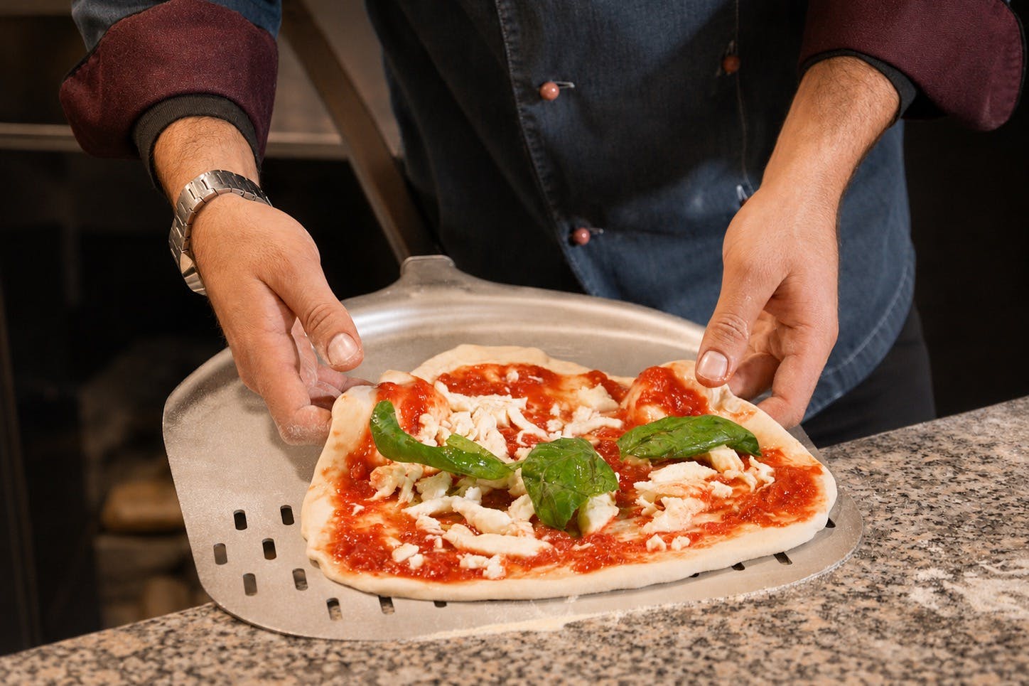 Una persona che tiene in mano una buccia di pizza con una pizza cruda condita con salsa di pomodoro, formaggio e foglie di basilico.