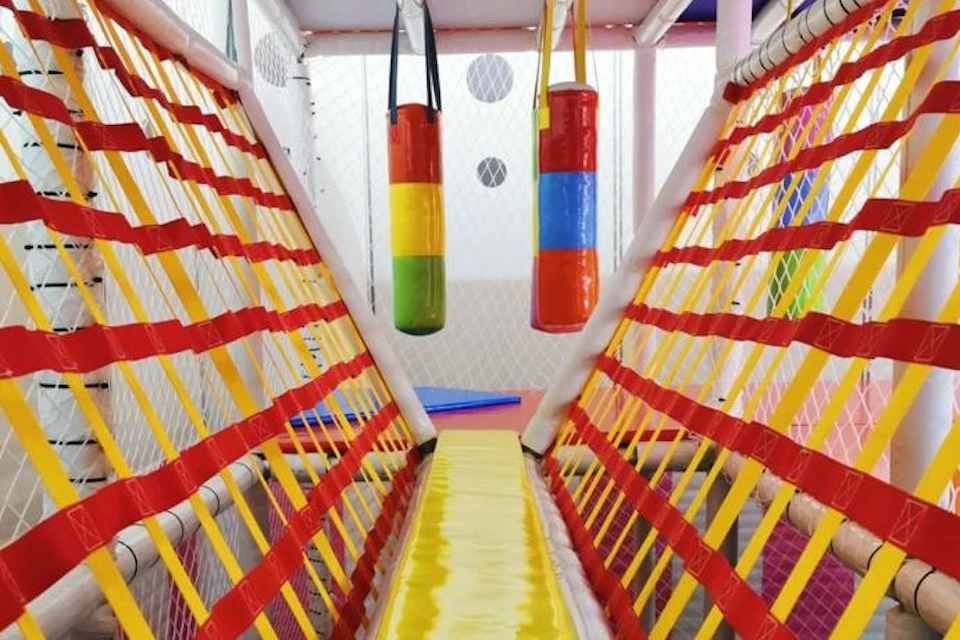 A colorful children's indoor play area with a narrow yellow path, red and yellow netting, and hanging padded cylinders.