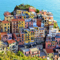 Kluster av färgglada byggnader i flera våningar på en klippa vid havet med utsikt över ett blått hav, med grönska i förgrunden.