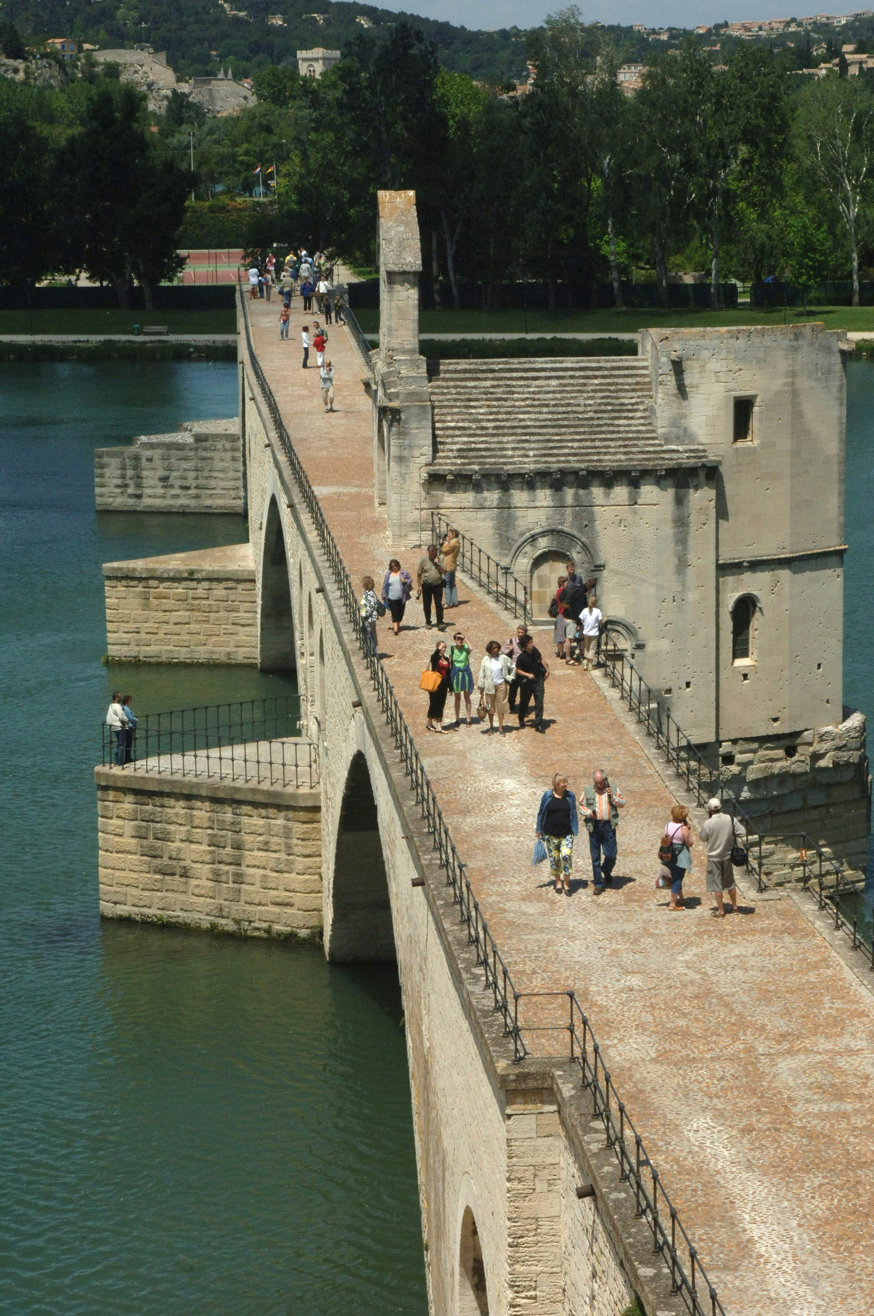 Lidé kráčející po historickém kamenném mostě přes řeku se stromy a budovami v pozadí.