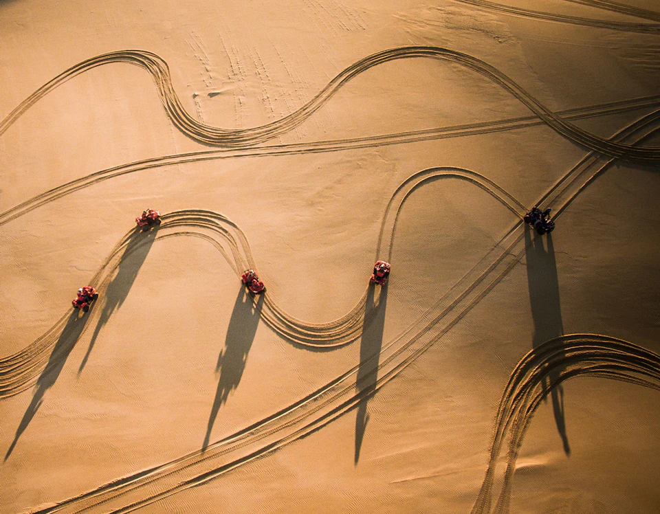 Aerial view of five ATVs making curved tracks in sandy terrain, casting long shadows in the sunlight.