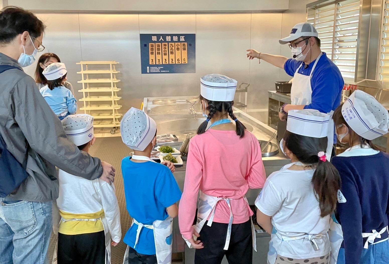 A group of children wearing chef hats and aprons listening to an instructor in a kitchen setting.