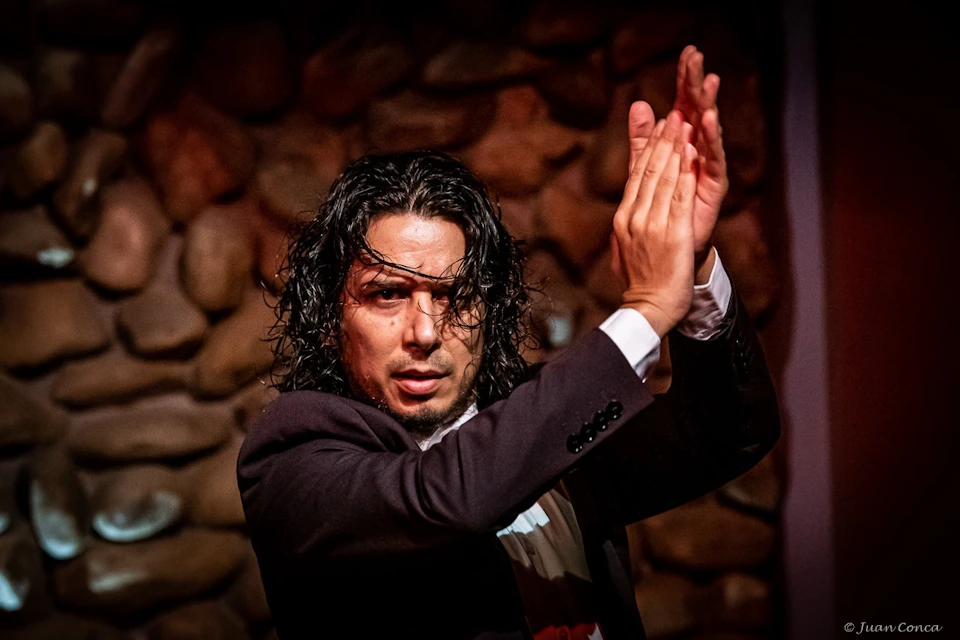 A person with wet hair clapping expressively in front of a wall made of round stones, wearing a dark suit.