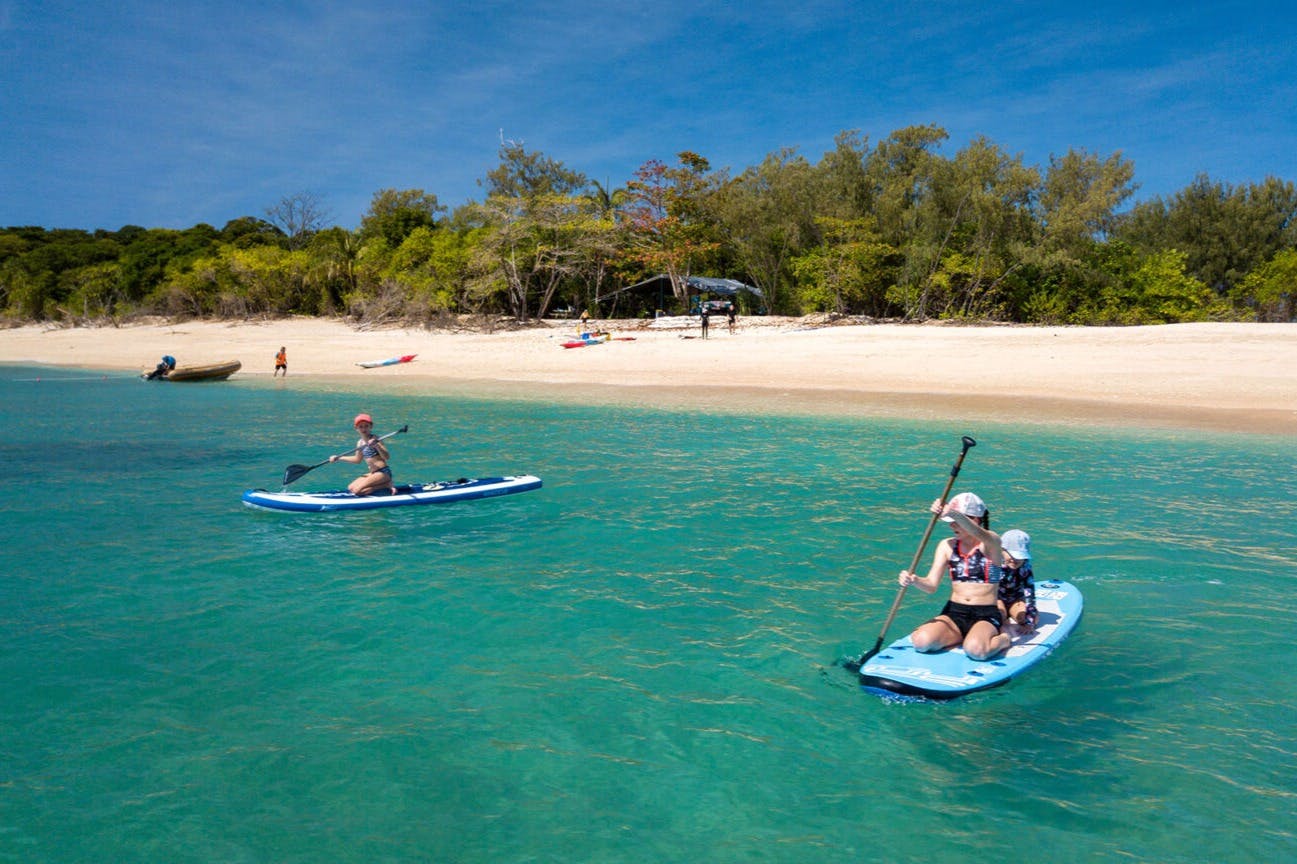 Lidé jezdí na paddleboardu na průzračné tyrkysové vodě poblíž písečné pláže se stromy a kajaky v pozadí.