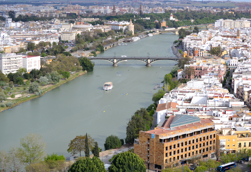 Aerial view of a river with a bridge spanning across, surrounded by buildings, trees, and several boats on the water.