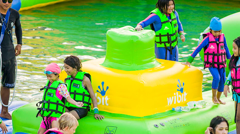 Children wearing life jackets play on a colorful inflatable water obstacle branded "Wibit."